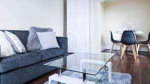 A modern living room with a grey sofa, a glass coffee table, and a dining area with a white table and black chairs.