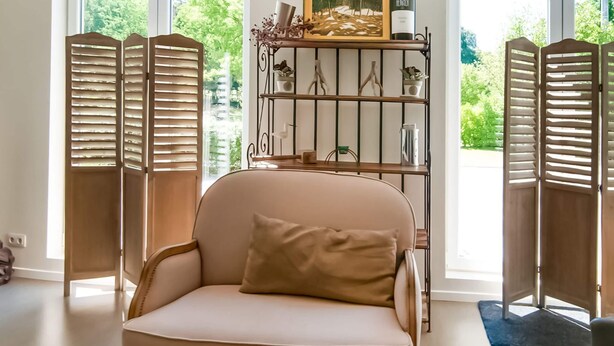 A room with a beige armchair, a metal shelf with a wine bottle, and a painting. - Enticing Holiday Home in Treignes near Forest (Viroinval)