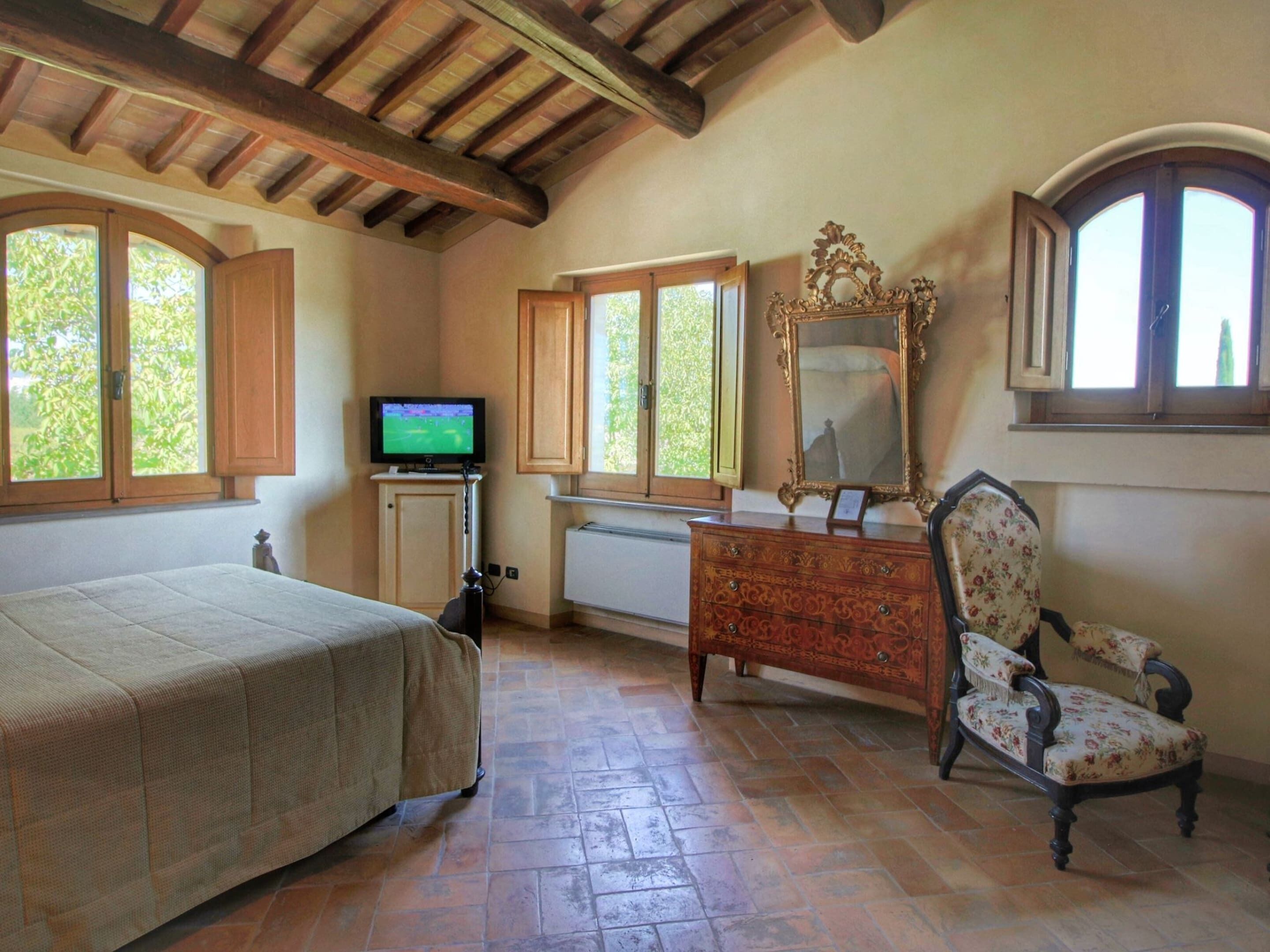 A bedroom with a bed, a dresser, a chair, a television, and large windows.