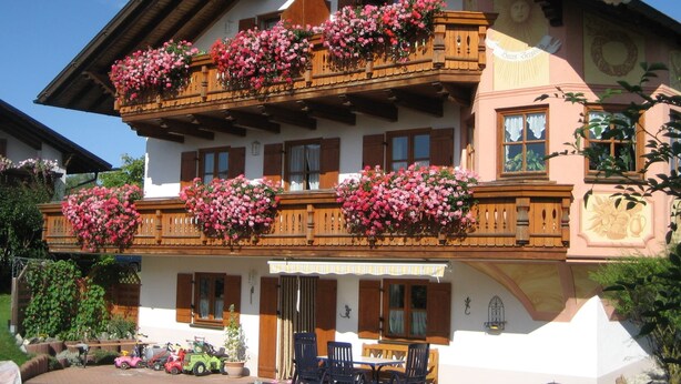 A traditional chalet with wooden balconies, a covered patio, and a garden area with toys and a lawnmower. - Apartment in Ingenried With Bavarian Alps View (Ingenried)