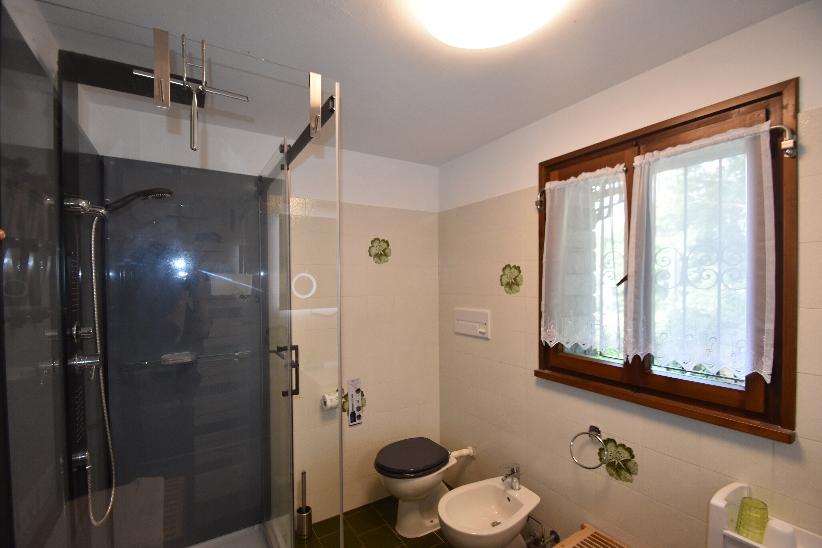 A bathroom with a glass shower enclosure, a toilet, a bidet, and a window with curtains.