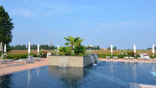 Una piscina con caída de agua, rodeada de sillones de descanso y sombrillas, con vista a un edificio lejano y campos abiertos.