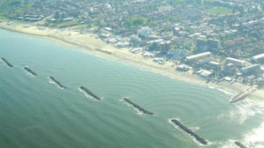 Vista aérea de un pueblo costero con playa, edificios y un muelle que se adentra en el agua.