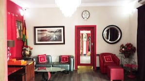 Uma sala de estar com um sofá vermelho, um sofá verde e uma poltrona vermelha. Há um lustre no teto, um relógio na parede e um espelho na parede.