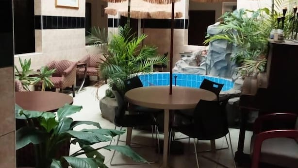 Um espaço amplo e interno, com uma mesa redonda e cadeiras, cercado por plantas em vasos.