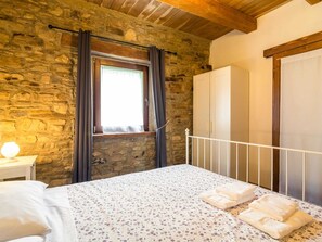 A bedroom with a stone wall, a wooden ceiling, a bed with a floral bedspread, a nightstand with a lamp, and a wardrobe. - Pretty Farmhouse With Pool (Sestino)
