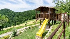 Une structure de jeu en bois avec un toboggan jaune, entourée d’une végétation luxuriante et de collines.