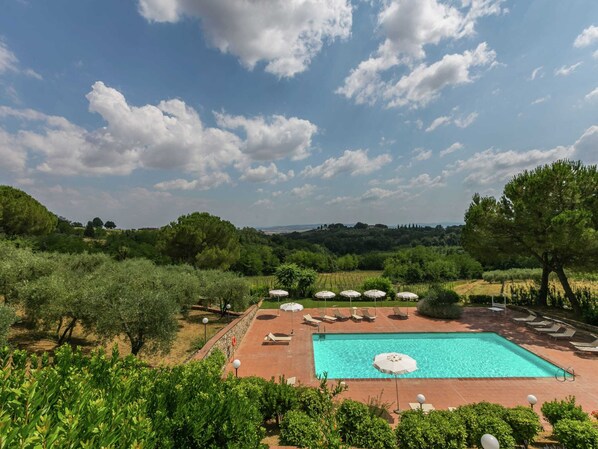 A rectangular swimming pool with a tiled deck, surrounded by lounge chairs and umbrellas, set against a backdrop of lush greenery and a clear sky.