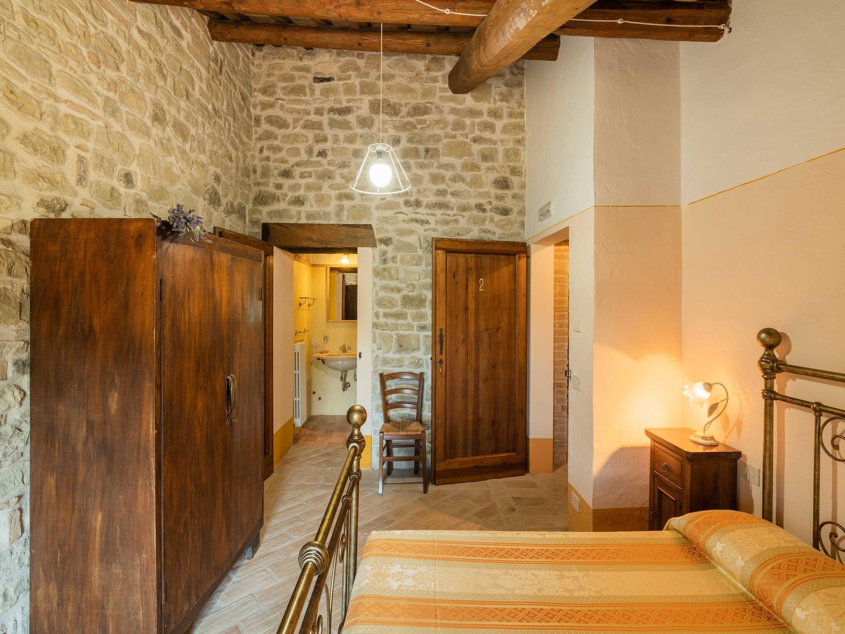 A rustic room with a stone wall, wooden wardrobe, a bed with patterned bedding, and a small bedside table with a lamp.