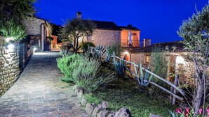 Ein gepflasterter Weg führt zu einem Steinhaus mit einem Balkon, umgeben von Grün und beleuchtet durch künstliches Licht.