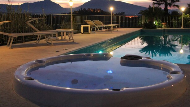A swimming pool with lounge chairs and a hot tub, surrounded by a fence and palm trees, with mountains in the background. - Mansion in Case Alte With Pool & Nature Trails (Sant'Omero)