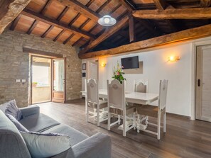 A spacious living area with a stone wall, wooden ceiling beams, a dining table with white chairs, and a grey sofa. - Tranquil Farmhouse in Sestino (Sestino)