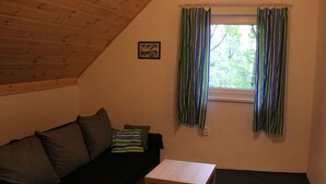 Et hyggeligt rum med skråvægge, træloft, en sort sofa med puder, et lille hvidt bord og et vindue med striber i gardinerne.