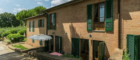 A two-story brick building with green shutters, a stone staircase, and a small patio area with a table and umbrella.