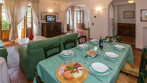 Un'area pranzo con un tavolo apparecchiato per quattro persone, una televisione sullo sfondo e vista sull'esterno attraverso ampie finestre.