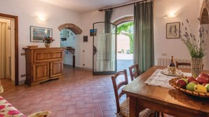 Comedor con mesa de madera, sillas y una cesta de frutas. Se ve una cocina a través de un arco, y una puerta que da al exterior.