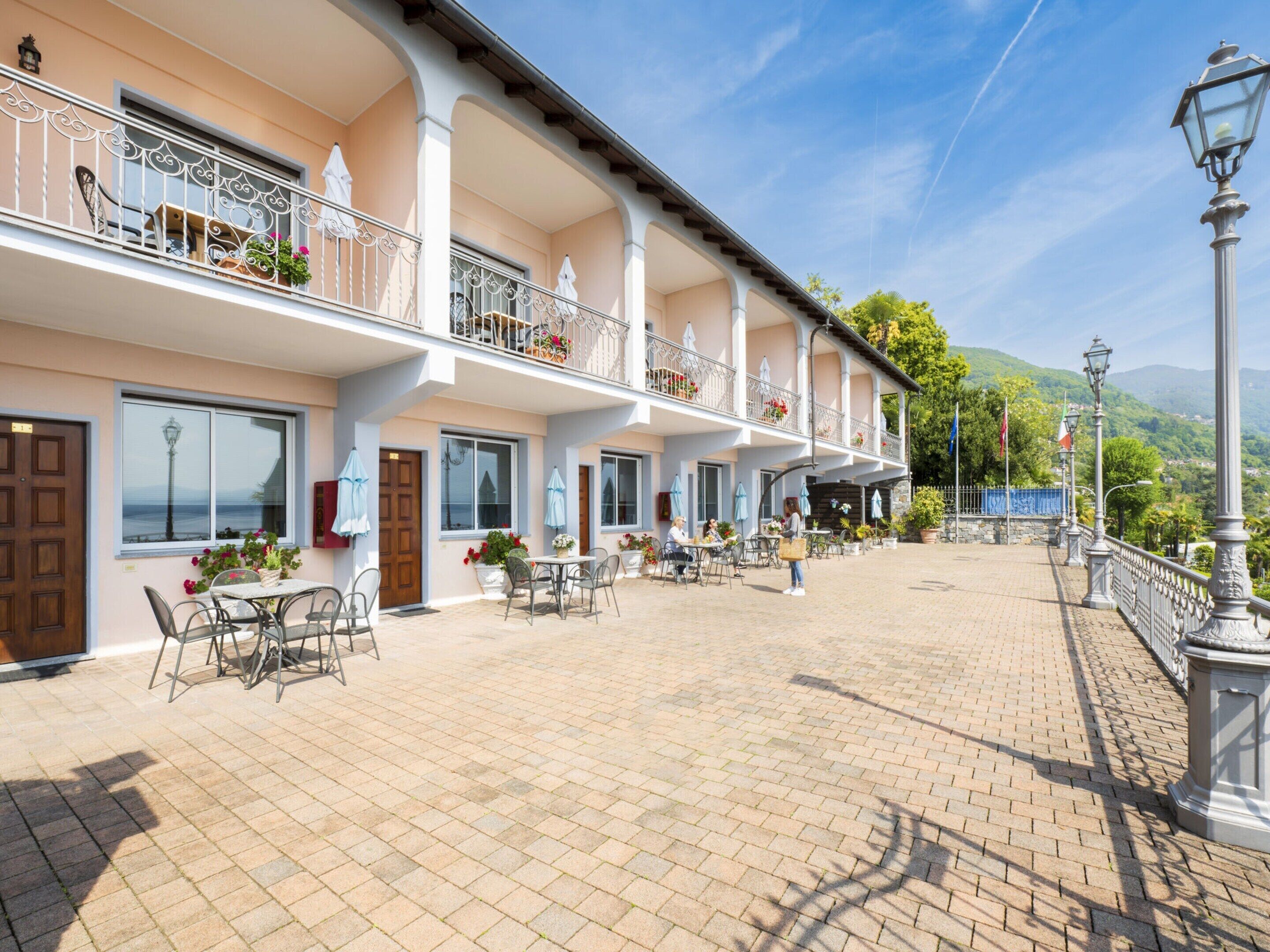 A hotel with outdoor seating, multiple balconies with flower boxes, and a paved courtyard.