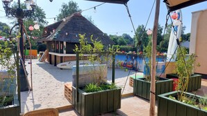 Een terras buiten met rieten stoelen en plantenbakken, een rieten dakconstructie en een zwembad op de achtergrond.