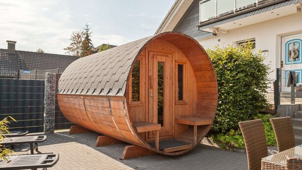 A wooden barrel-shaped structure with a door and windows, situated on a paved patio with outdoor furniture. - Entspannender Ruckzugsort in Langewiese (Winterberg)