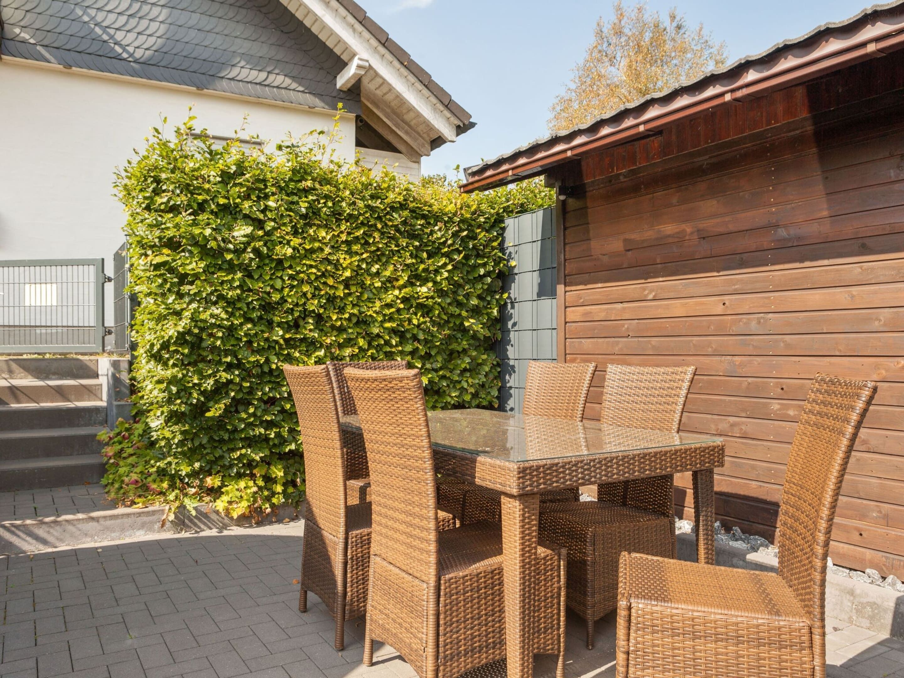 An outdoor patio with wicker furniture and a glass table, surrounded by a hedge and a wooden building.