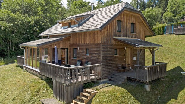 A wooden cabin with a covered porch and a small deck, surrounded by a grassy area and trees.