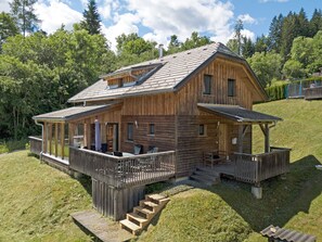 Een houten hutje met een overdekte veranda en een klein terras, omringd door een gazon en bomen.