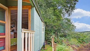 A wooden house with a porch, a visible kitchen area, and a lush green landscape in the background.