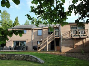 A two-story wooden house with a stone retaining wall, outdoor seating, and a staircase leading to a balcony.