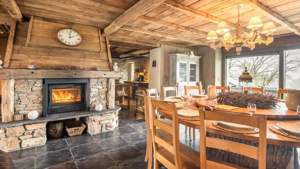 Ein rustikaler Innenraum mit Kamin aus Stein, Holzbalken an der Decke und einem Essbereich mit Holzsesseln sowie einem für vier Personen gedecktem Tisch.