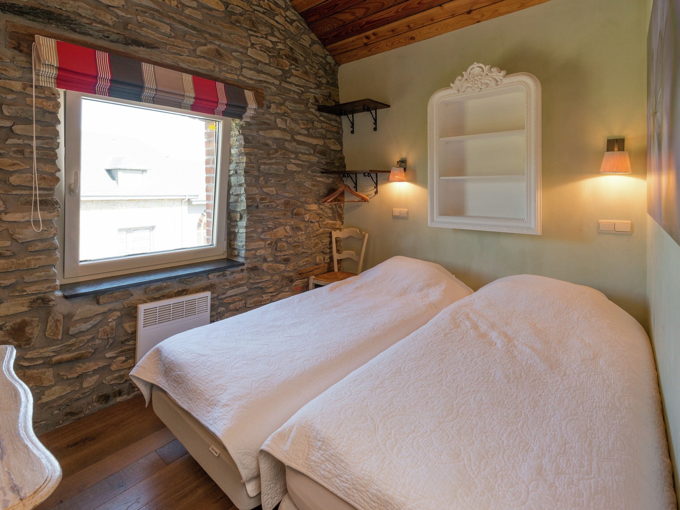 Ein Schlafzimmer mit zwei Betten, Steinwänden, einem Fenster mit Vorhängen, einem Sessel und Wandleuchten.