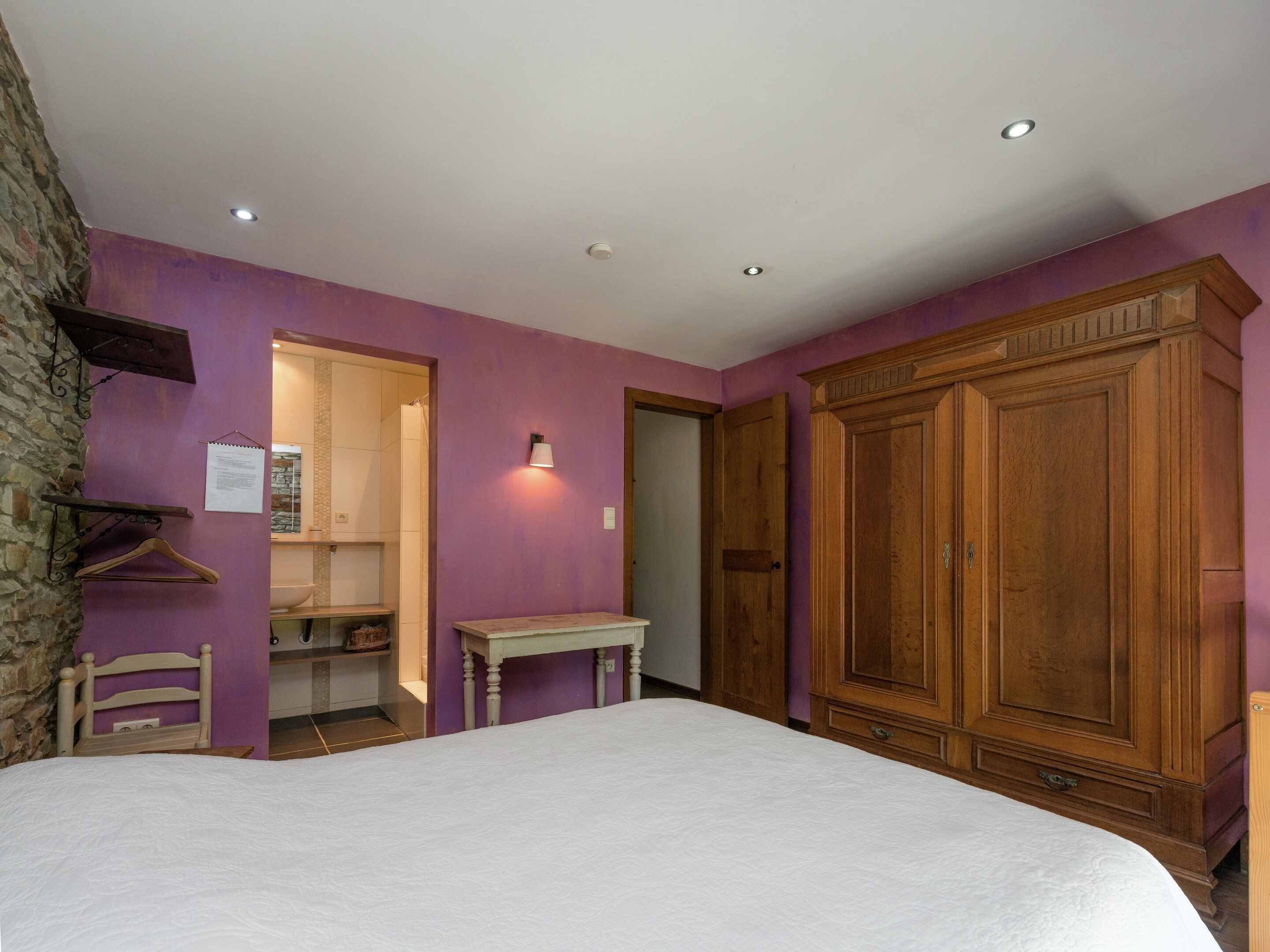 A bedroom with a bed, wooden wardrobe, a small table, and a bathroom visible through an open door.