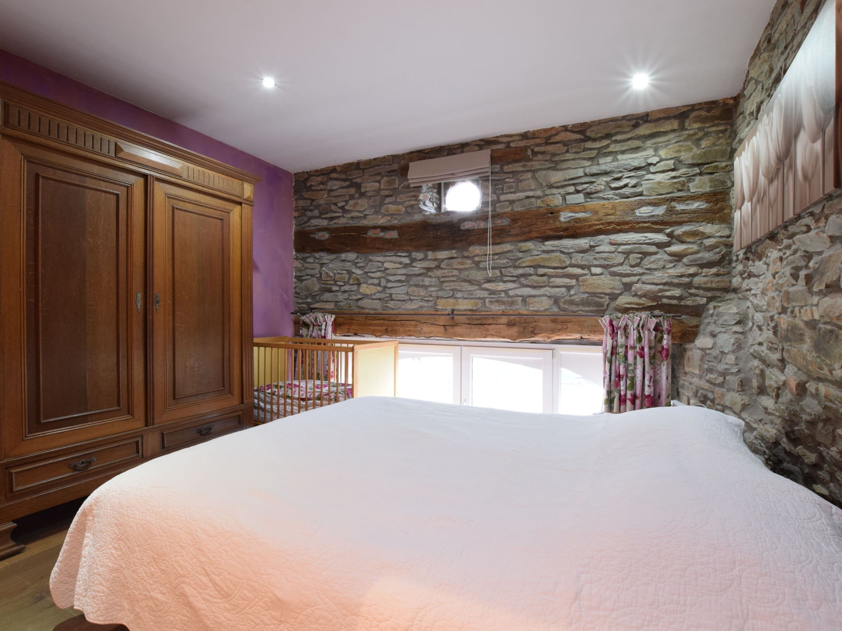 A bedroom with a large bed, wooden wardrobe, stone wall, and a small balcony with a view.