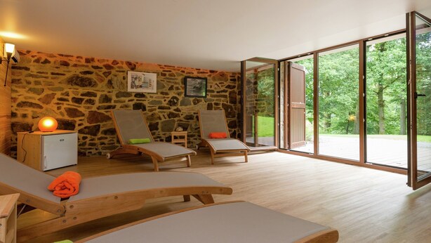 A room with stone walls, two wooden lounge chairs, a small table, and a large sliding glass door leading to an outdoor area. - Tenneville Villa With Sauna (Tenneville)