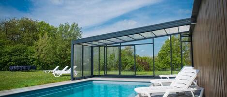 A swimming pool with clear blue water, surrounded by white lounge chairs and a glass-enclosed patio area.