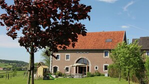 A large brick building with a red-tiled roof, a central arched entrance, and a small adjacent structure with a green roof. - Holiday Home in Limbourg With Garden (Limbourg)