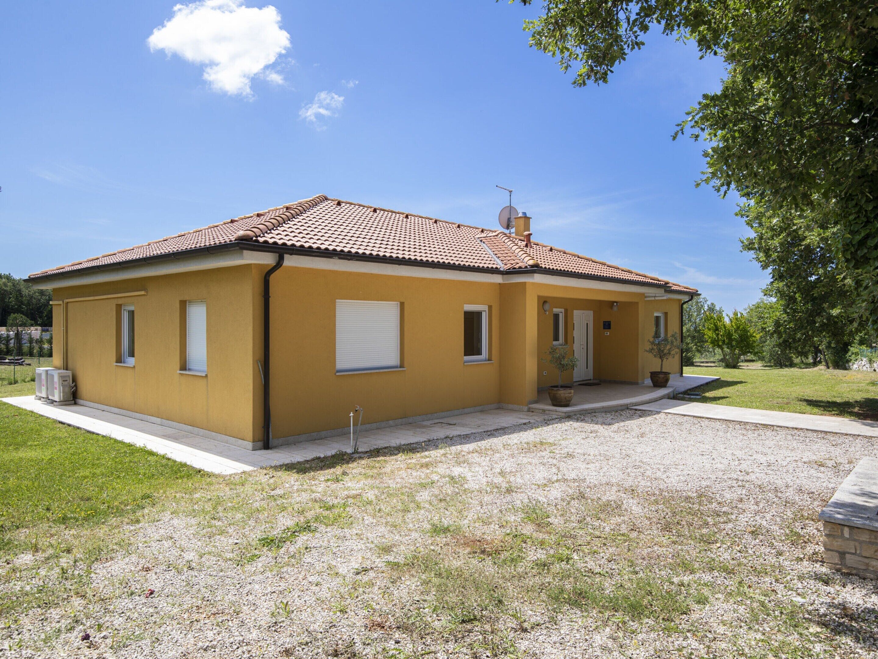 A single-story yellow building with a tiled roof, a small porch, and a gravel driveway.