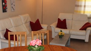 A living room with two white leather sofas, each with a red pillow, a wooden coffee table with a vase of flowers, and a wooden dining table in the foreground. - Chicke Ferienwohnung in Struppen (Struppen)