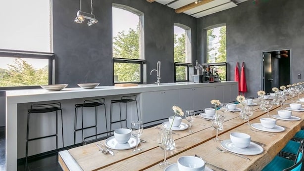 A modern dining area with a long wooden table set for a meal, large windows, and a kitchen area visible in the background. - Holiday Home in Denee for 21 People (Anhée)