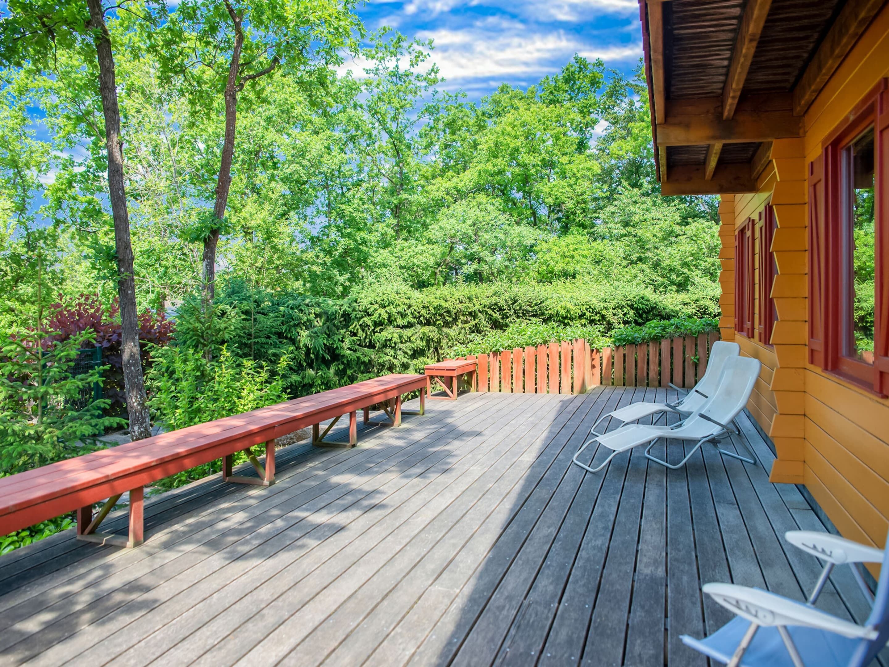 A wooden deck with benches and lounge chairs, surrounded by lush greenery and trees.