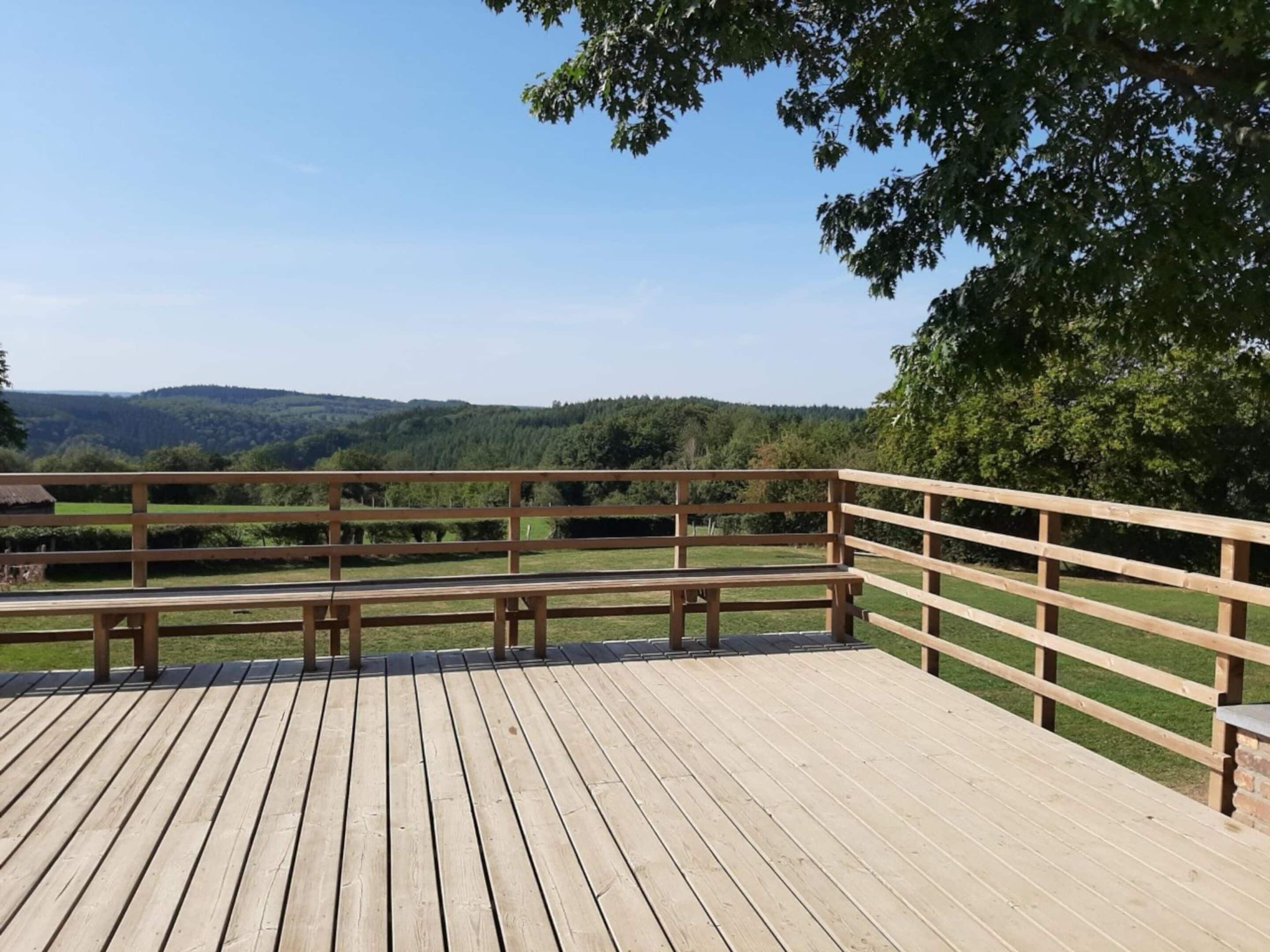 A wooden deck with benches and a railing, overlooking a grassy field and forested hills.