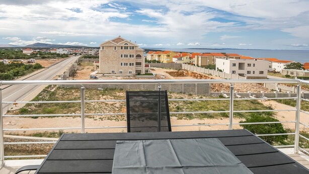 A balcony with a view of a coastal area, featuring a building with a stone facade and a modern structure with orange roofs. - Apartment in Novalja Near Zrce Beach (Novalja)