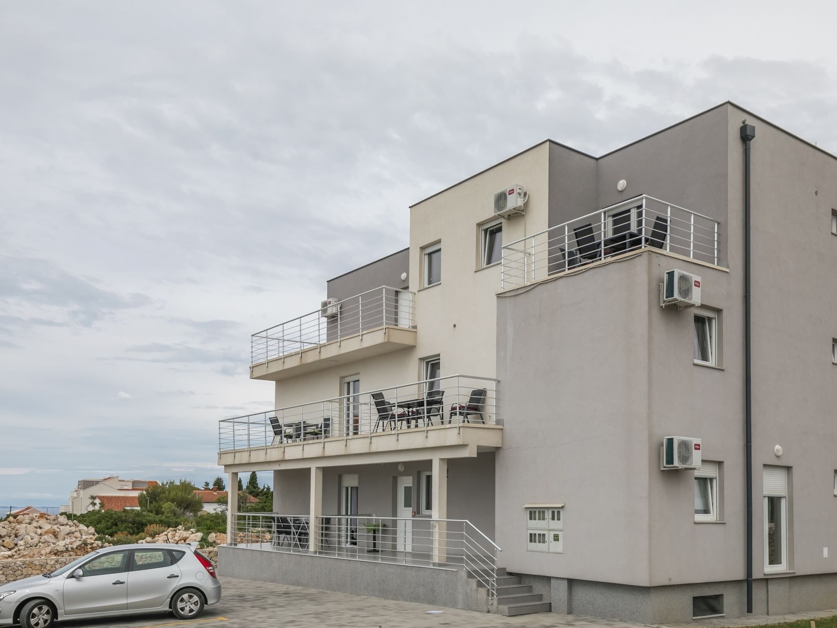 A modern two-story building with balconies, a car parked in front, and a stone wall in the background.