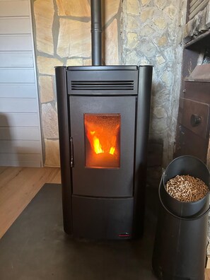 A wood-burning stove with visible flames, a stone wall, and a metal container of wood pellets. - Chalet in Guntersberge Near Ski Trails (Harzgerode)