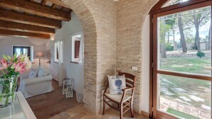 Une pièce chaleureuse avec un mur en pierre, des poutres apparentes, une grande fenêtre, un fauteuil et un canapé blanc.