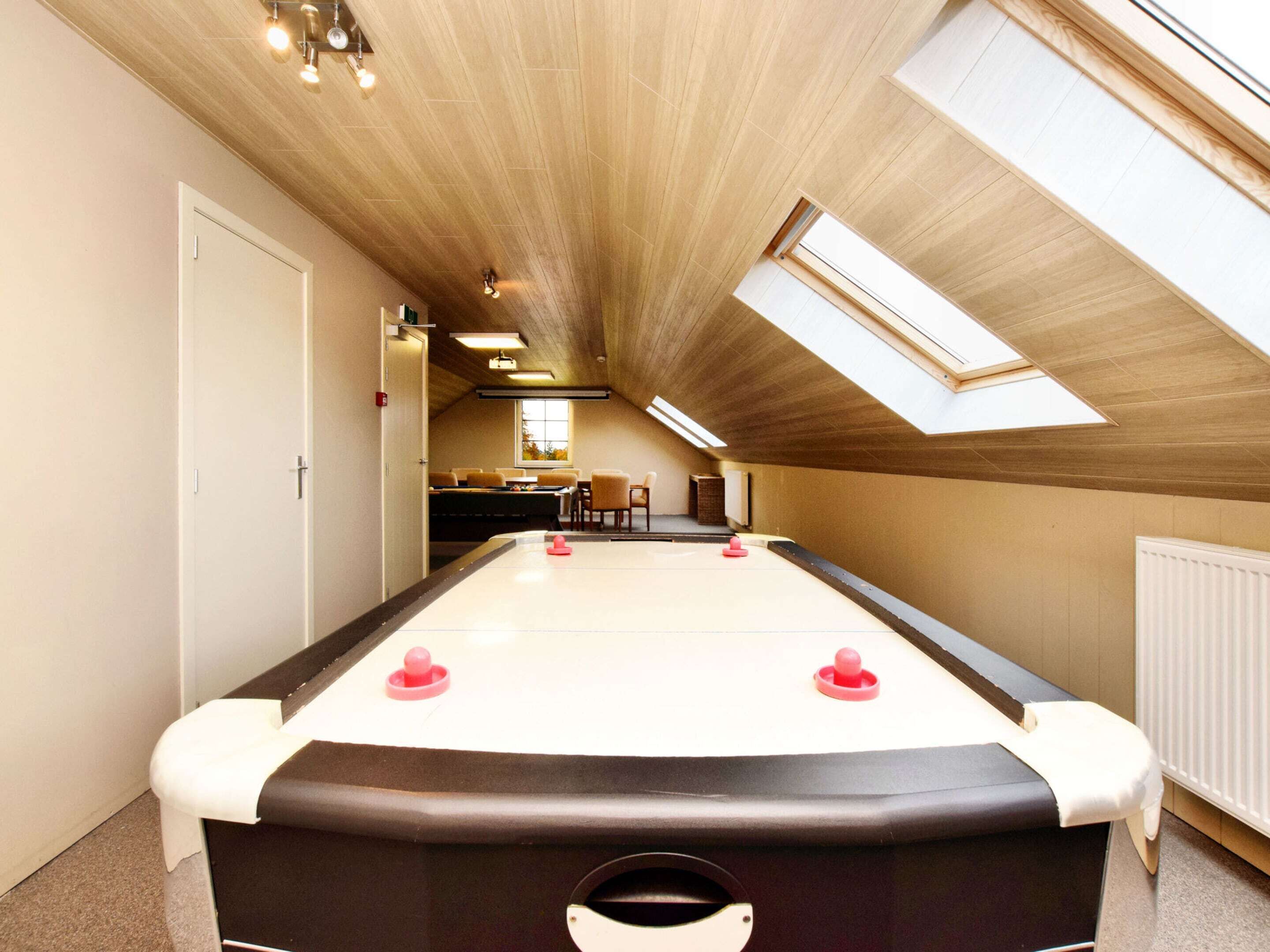 Uma mesa de bilhar em um espaço com teto inclinado e claraboias, uma porta à esquerda e uma pequena área de refeições ao fundo.