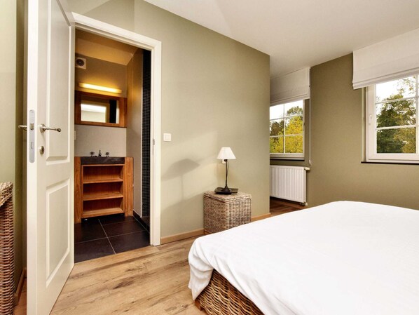 Une chambre à coucher avec un lit, une table de chevet avec une lampe, une fenêtre donnant sur des arbres, et une salle de bain avec un miroir et un lavabo.