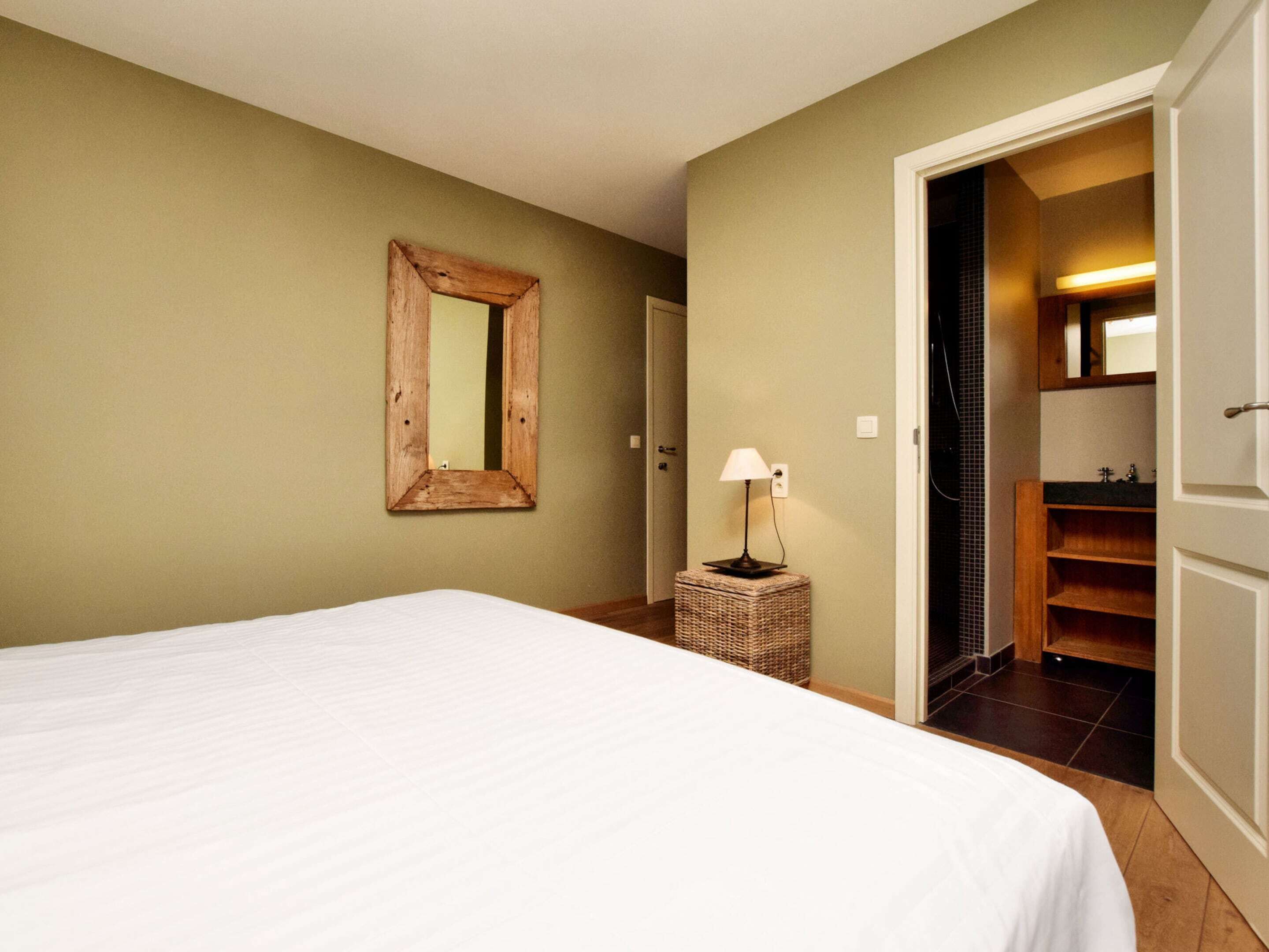 A bedroom with a large bed, a wooden-framed mirror, a bedside lamp, and a wicker side table.