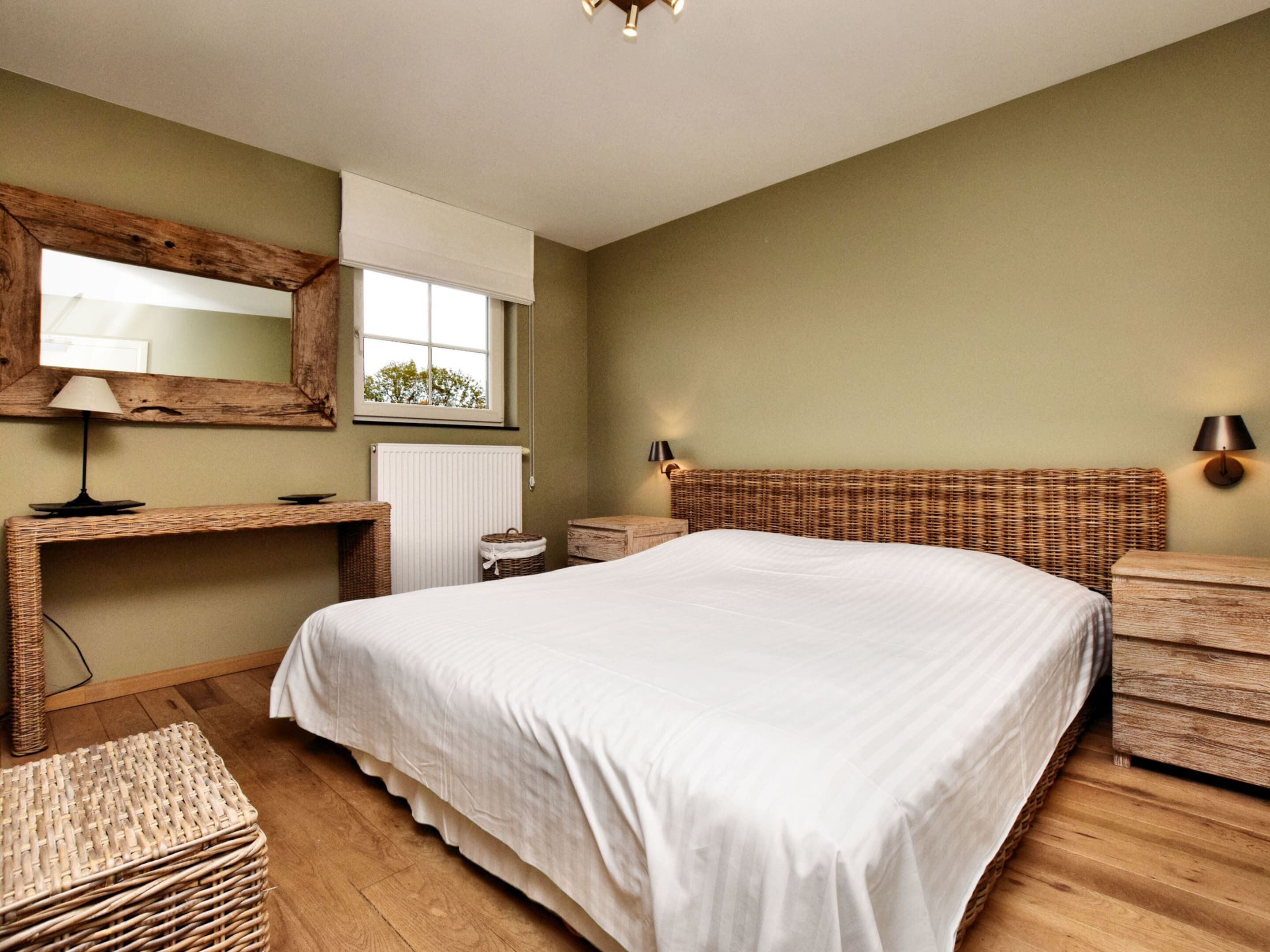 Une chambre avec un grand lit, une tête de lit en bois, une table de chevet, une lampe, un miroir, une fenêtre donnant sur un beau paysage et un radiateur.
