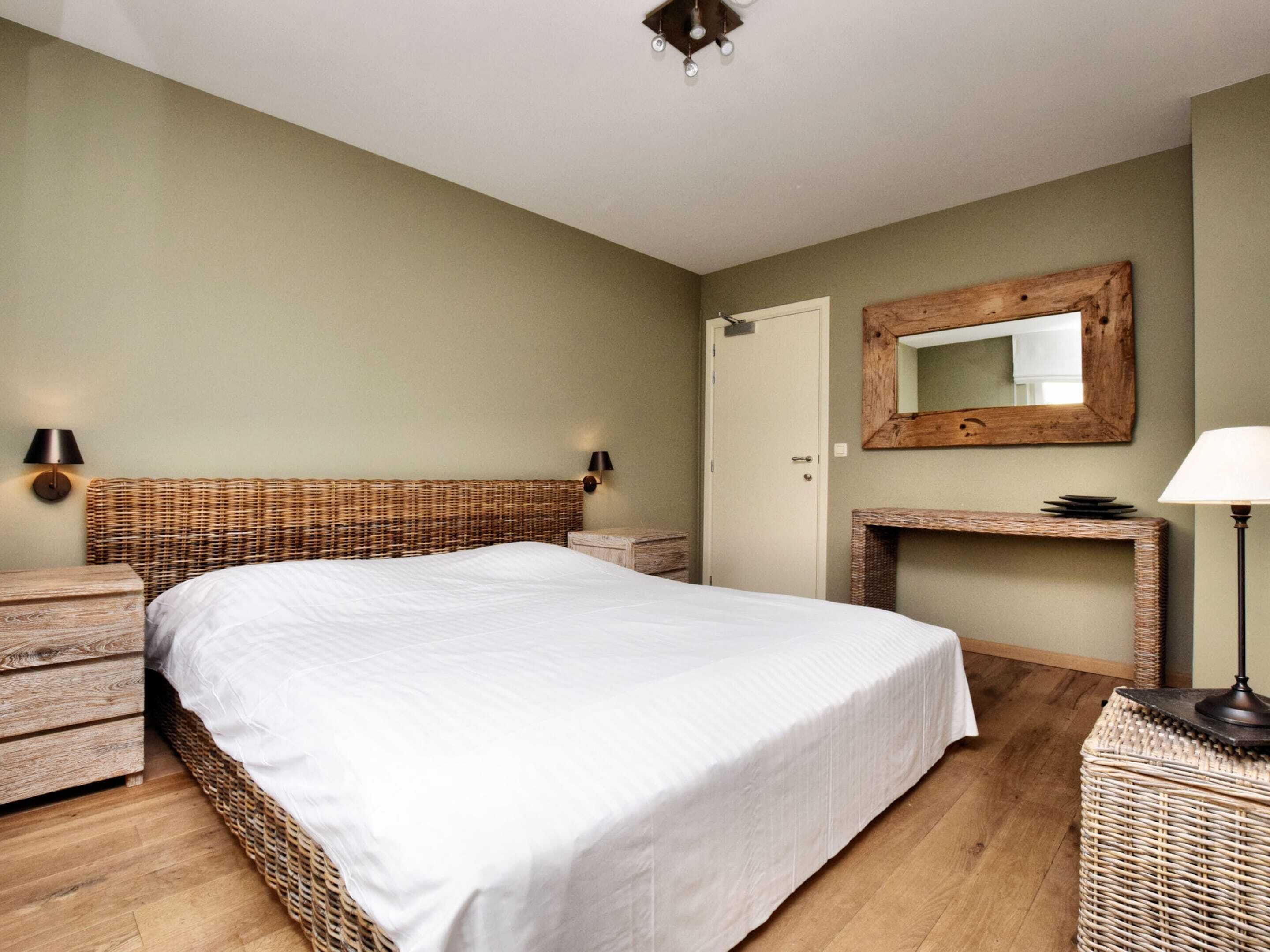 A bedroom with a large bed, a wooden headboard, a nightstand, a bedside table, a mirror, and a lamp.