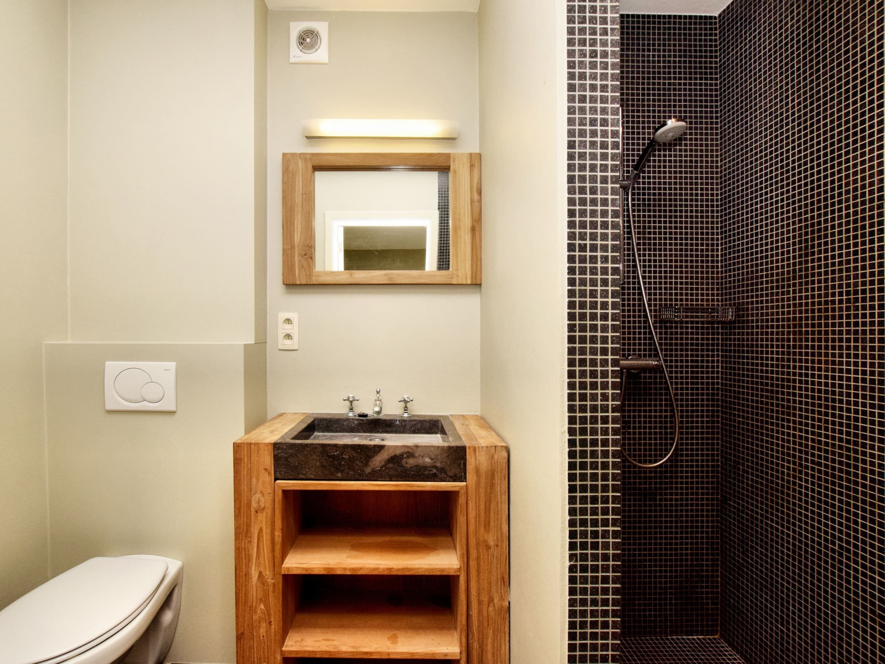 A bathroom with a wooden vanity, a toilet, a mirror, and a shower with tiled walls.